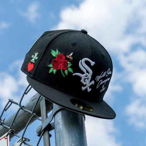 Black fitted cap with embroidered roses and logos, showcasing streetwear style against a blue sky.